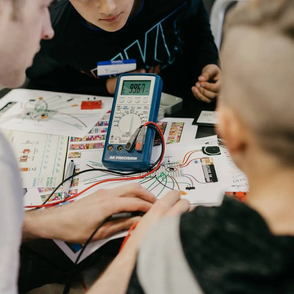 Students using a BK Precision multimeter to test circuit boards and wires on a cluttered workbench.