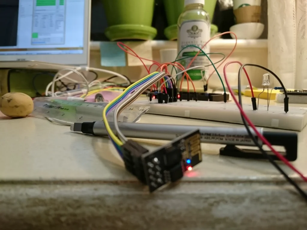 Electronics workbench with breadboard, ESP32 board, wires, potato, computer monitor, and kombucha bottle.