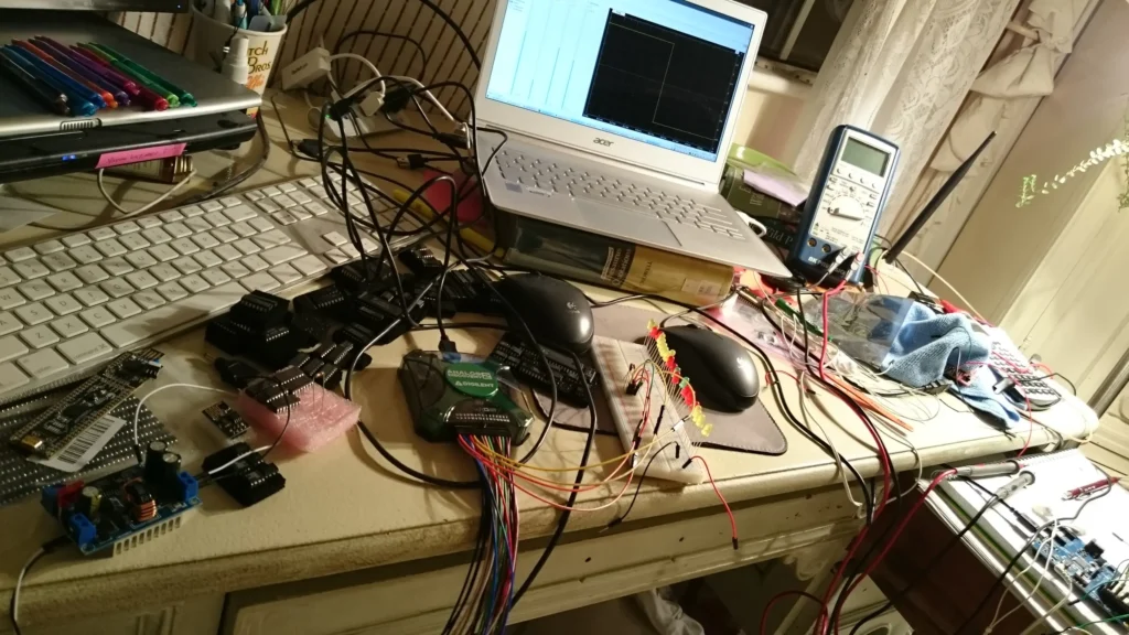 Cluttered electronics workbench with laptop, oscilloscope, multimeter, breadboards, wires, and Arduino components.