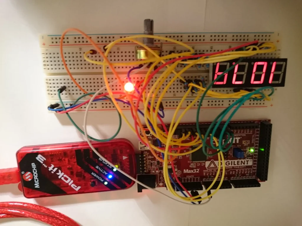 Breadboard circuit with multicolored wires connecting red 7-segment display showing "5001", potentiometer, red LED, USB programmer, and Xilinx Max32 FPGA board.