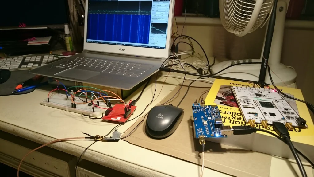 Laptop displaying oscilloscope waveforms beside breadboard circuit, Arduino, RF modules, and electronics book on a desk with fan.
