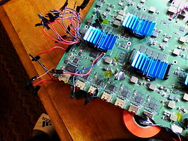 Close-up of green circuit board with blue heatsinks, colorful wires, tape, and Adidas shoe on wooden table.