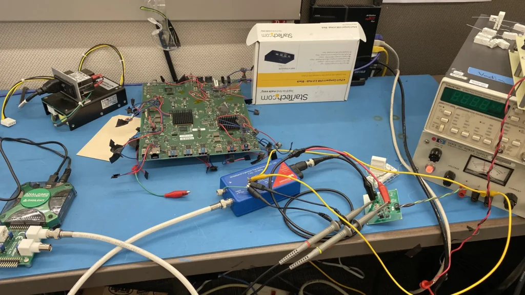Cluttered electronics workbench with disassembled circuit boards, oscilloscope, power supply, and tangled test leads on blue mat.