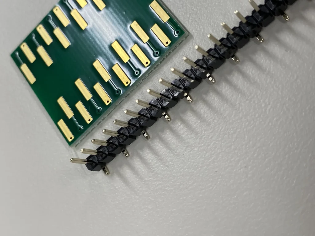 Green PCB with gold-plated edge contacts next to a black pin header connector on a white surface.