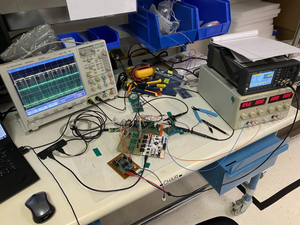 Electronics workbench with oscilloscopes, multimeter, circuit boards, tools, and laptop amid tangled wires.