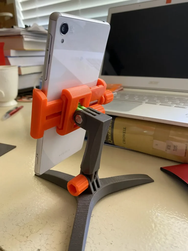 Orange and gray 3D-printed tripod phone stand holding a white Sony Xperia smartphone on a desk.