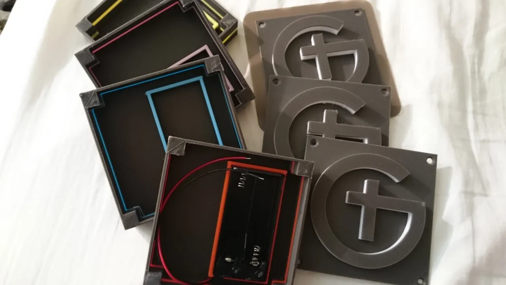 Assortment of colorful-edged black frames and gray plaques with raised G-cross logos, one containing a battery holder with wires.