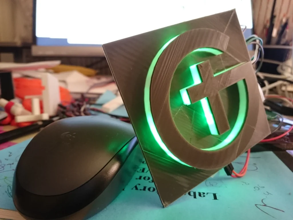 Illuminated 3D-printed green-glowing G with cross emblem on a desk beside Logitech mouse.