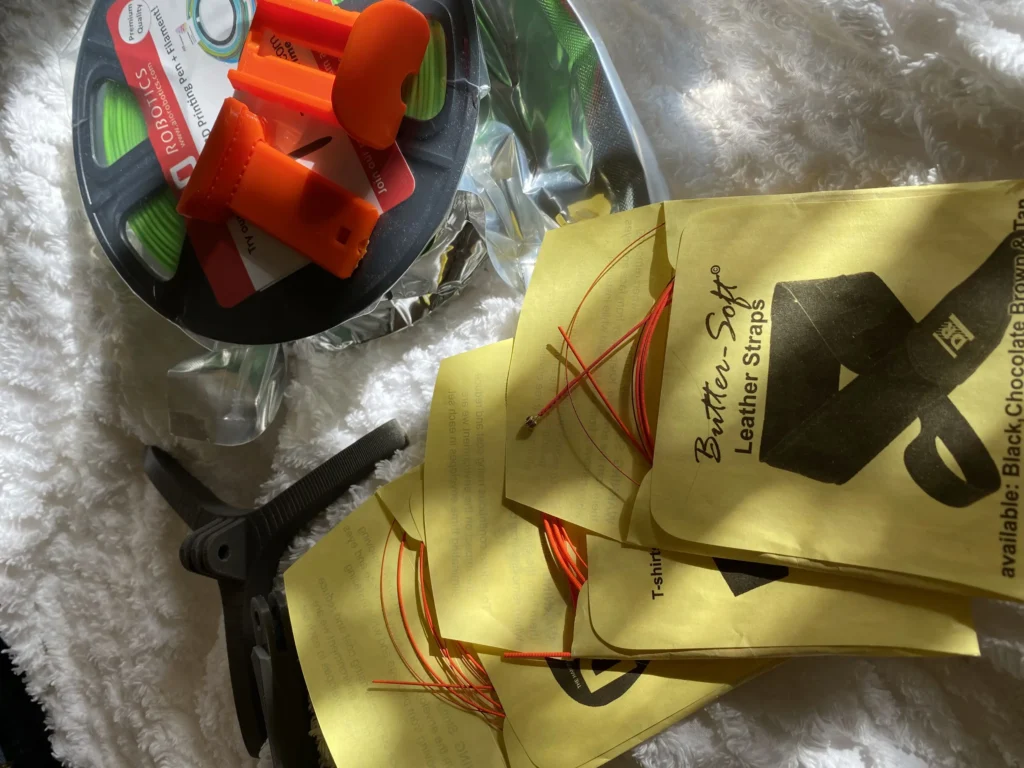3D printing filament spool, orange tools, red and orange wires in Butter Leather Straps packets, and pliers on fluffy white fabric.