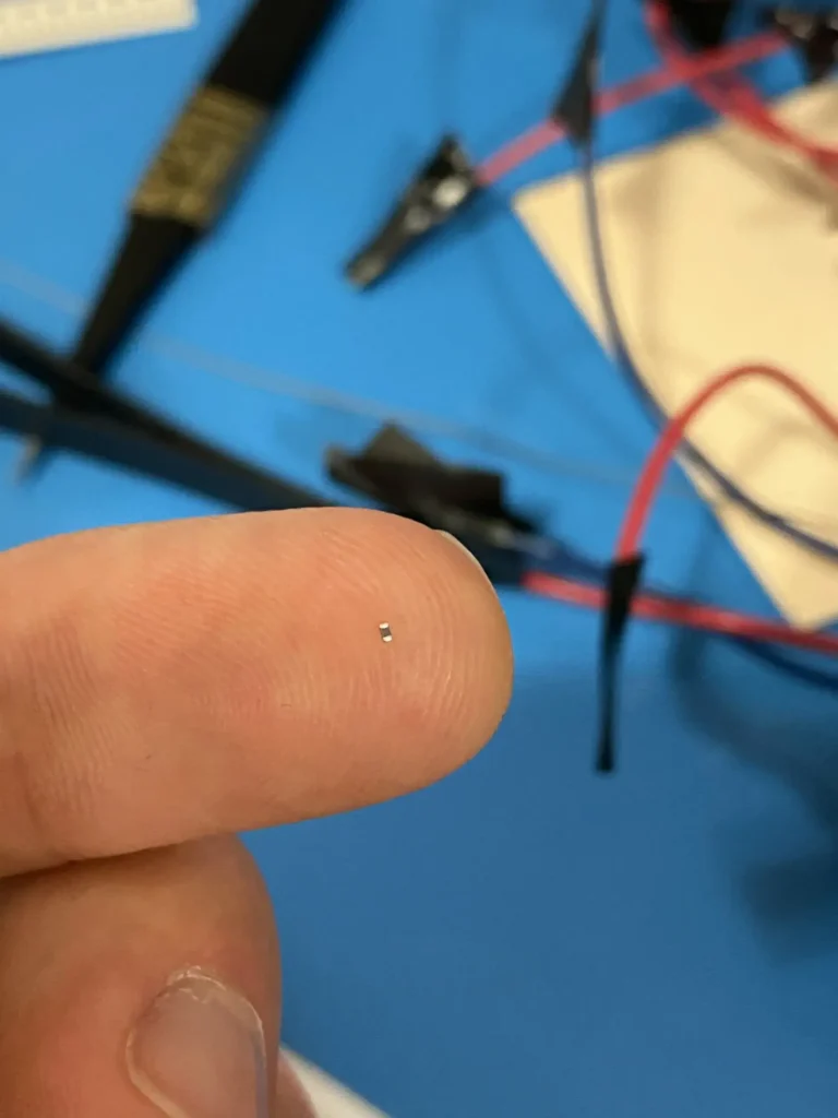 Tiny SMD resistor on fingertip amid electronics workbench tools and wires.