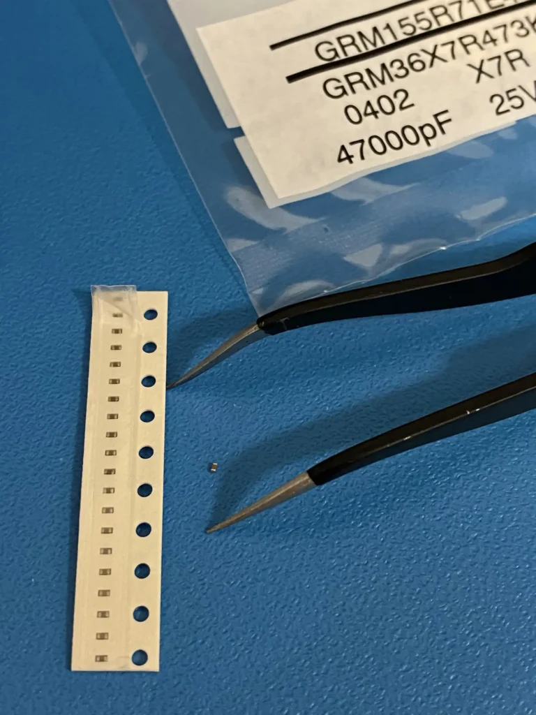 Tweezers holding tiny GRM155R71H473KA01D and GRM36X7R 0402 4700pF 25V capacitors next to SMD strip on blue mat.