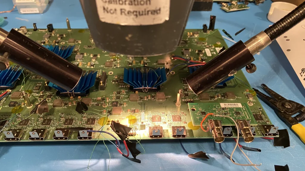 Close-up of green circuit board with blue heatsinks under hot air rework stations on workbench.