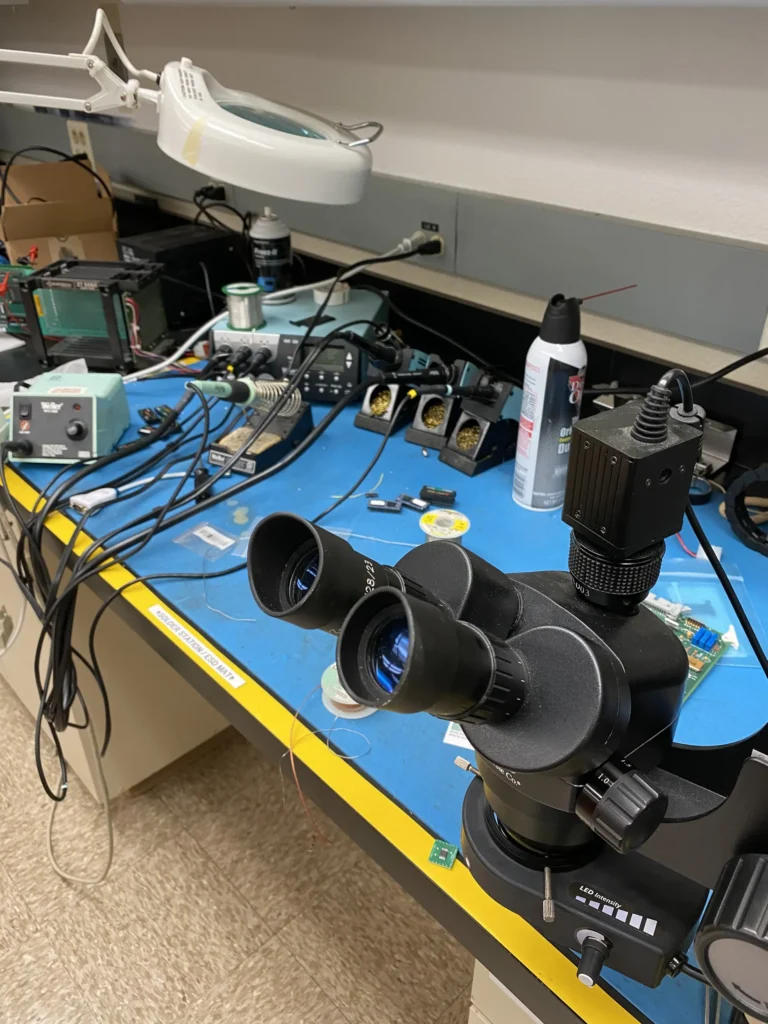 Cluttered electronics workbench with stereo microscope, soldering tools, components, and magnifier lamp.