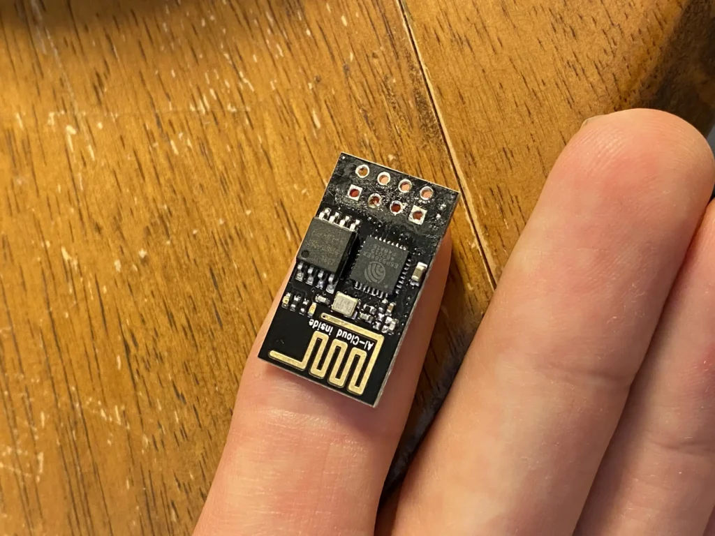 Tiny ESP32 LoRa module held on fingertip atop wooden table.