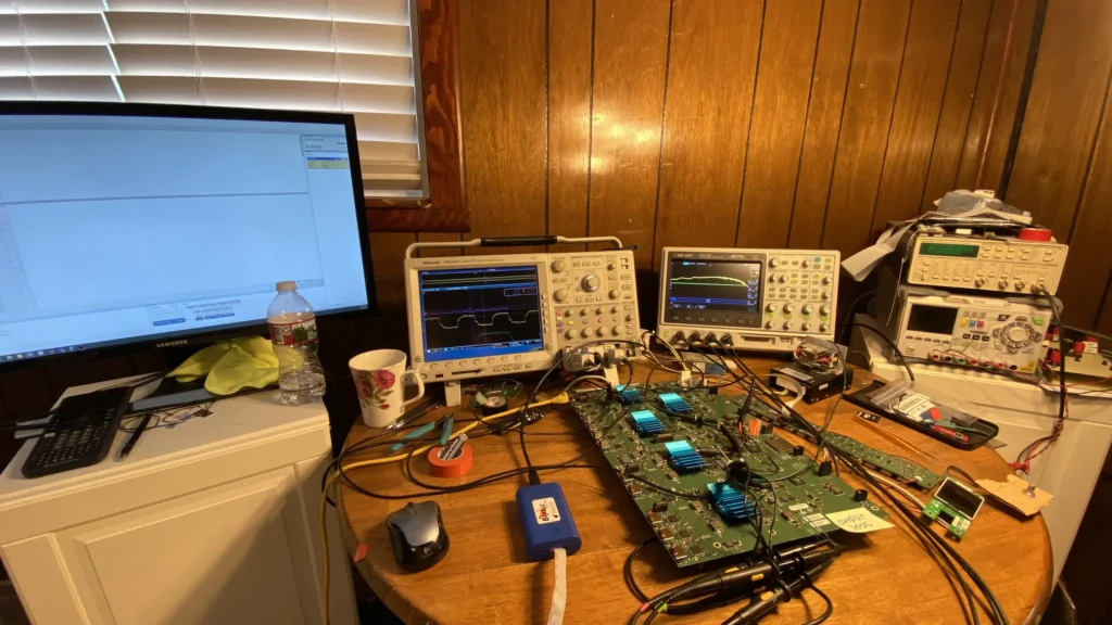 Electronics workbench with oscilloscopes, multimeters, computer monitor, circuit boards, and tools on a wooden desk.
