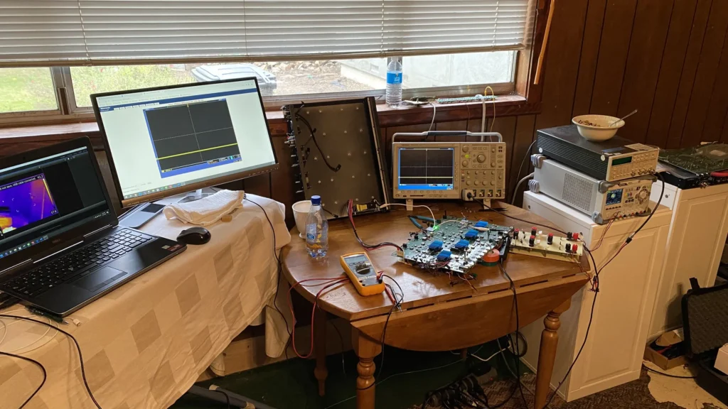 Electronics workbench with laptops, oscilloscopes, multimeter, circuit boards, and thermal camera on wooden table by window.