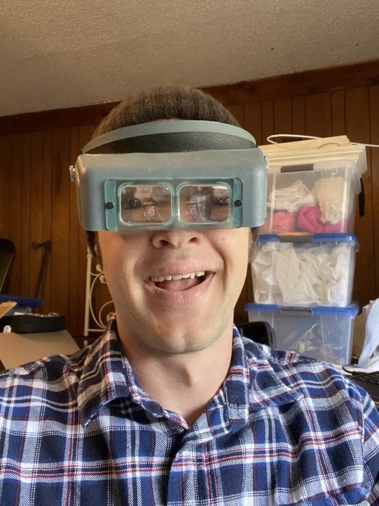 Man smiling while wearing gray vintage stereo viewer headset in cluttered workshop.