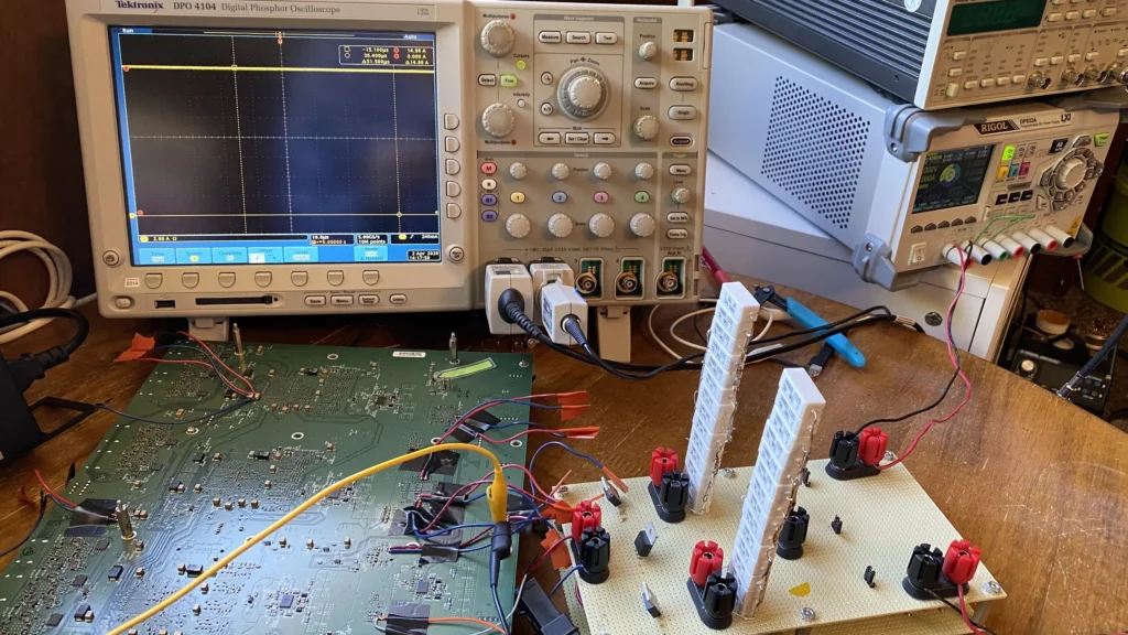 Electronics workbench with Tektronix oscilloscope, power supplies, green PCB, and LEGO-brick circuit prototype connected by wires.