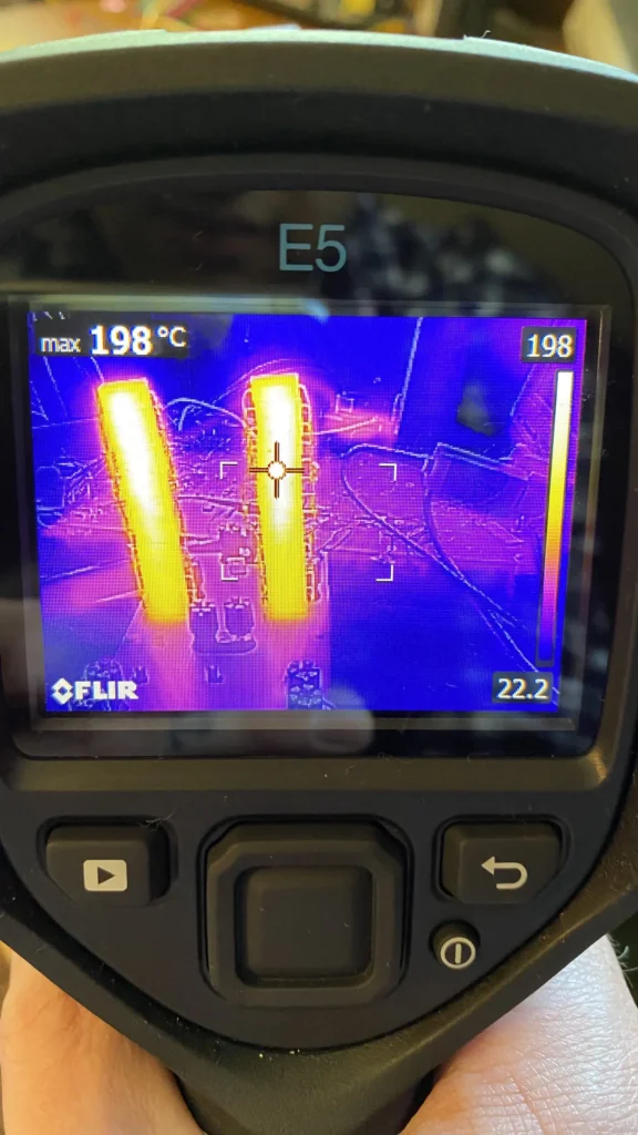 FLIR E5 thermal camera held in hand displaying hot yellow pipes amid purple cityscape at max 198°C.