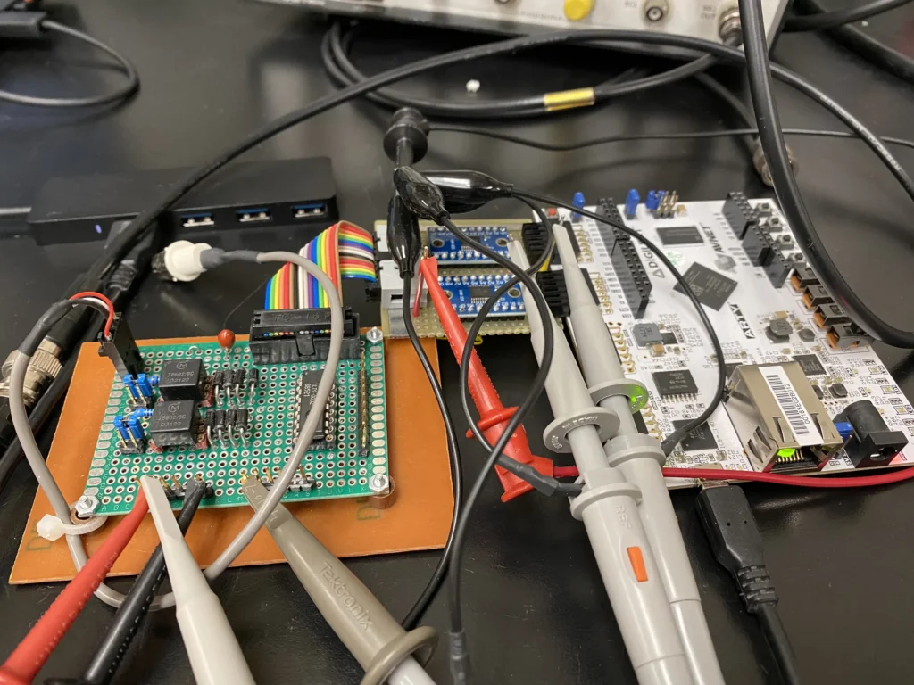 Prototype circuit board and microcontroller development board probed with multimeter leads on workbench.