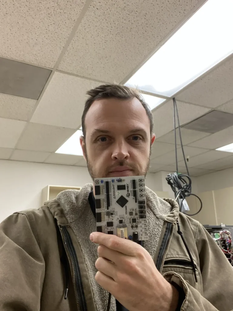 Man in hoodie holding Digilent Arty S7 FPGA board in office lab.