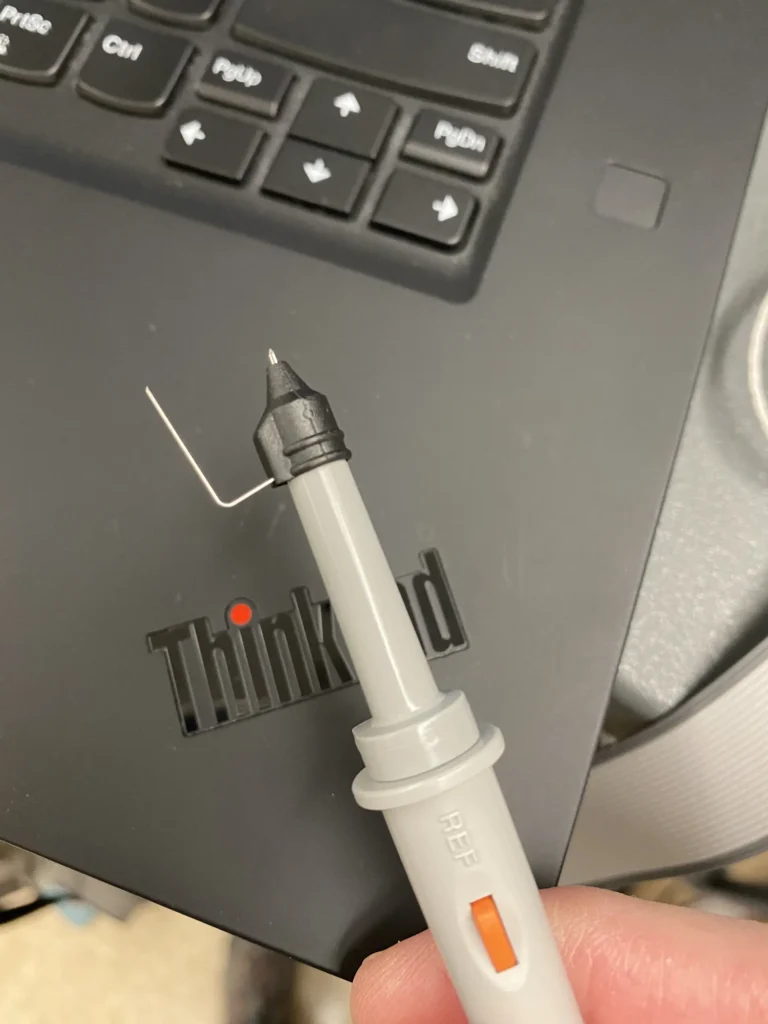 Hand holding gray REF multimeter probe in front of ThinkPad laptop keyboard.