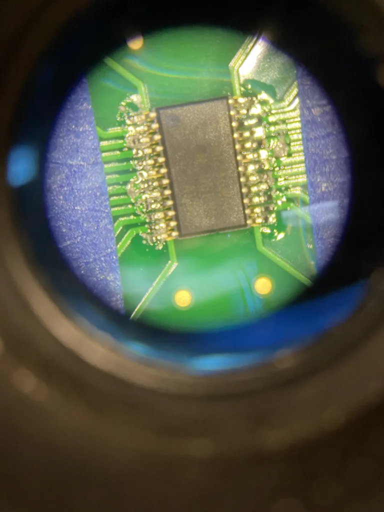 Close-up view through magnifying lens of green PCB with central black IC chip and gold pins.