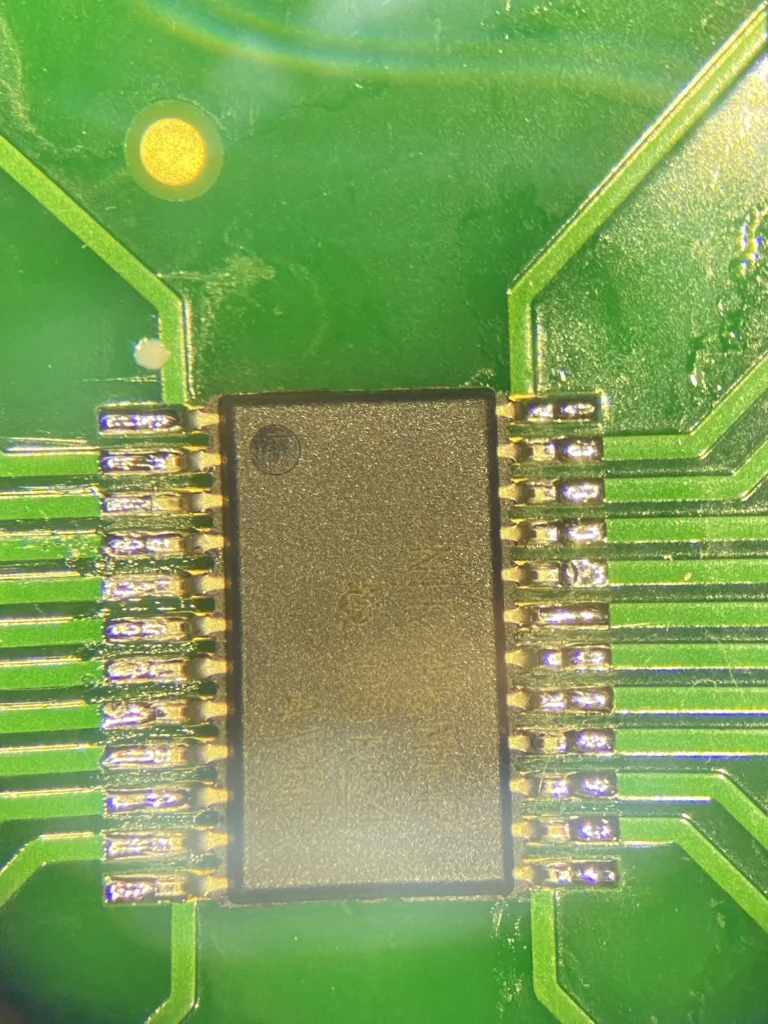 Close-up of a black SOIC integrated circuit soldered on a green PCB with gold traces and a yellow LED nearby.