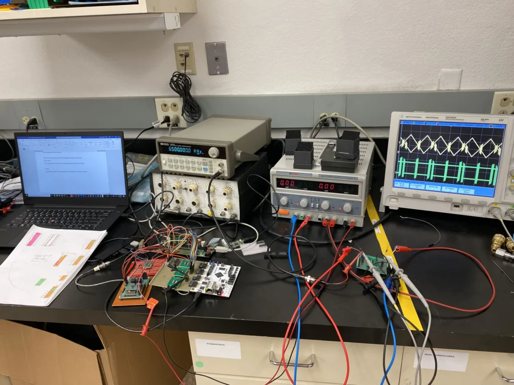 Electronics workbench with oscilloscopes, multimeters, power supplies, laptop, circuit boards, and tangled wires.