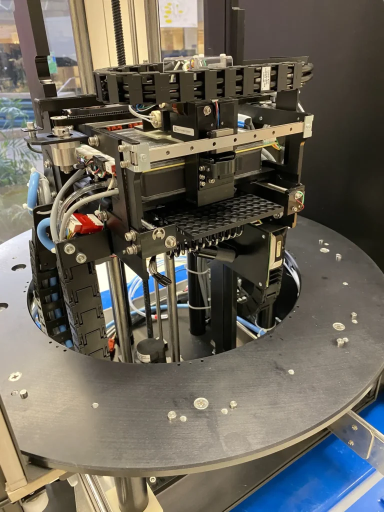 Close-up of a robotic automation machine with black frame, multiple rails, motors, and wiring on a circular platform.