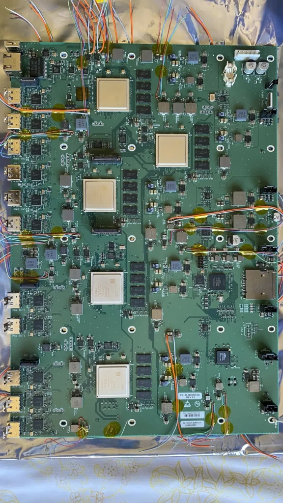 Green circuit board with multiple HDMI ports, gold heatsinks, capacitors, and multicolored wiring on foil background.
