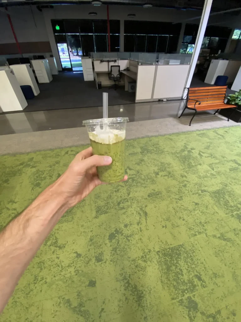 Hand holding green smoothie with whipped cream in modern office lounge with cubicles and green carpet.
