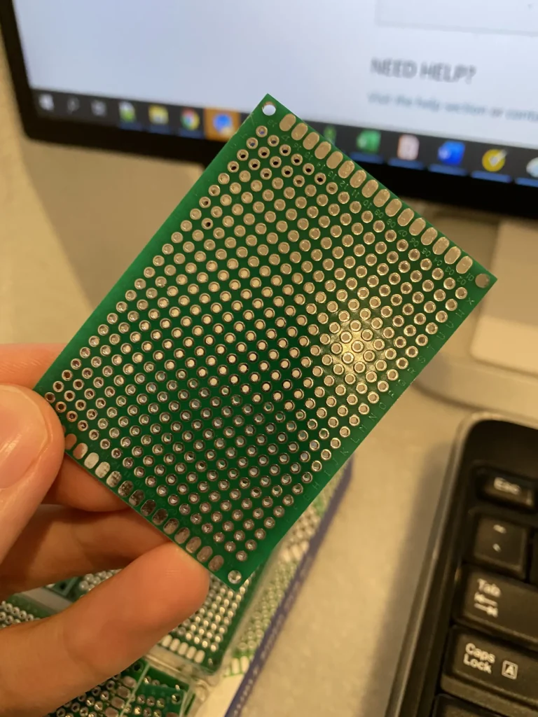 Hand holding green perforated prototyping PCB near computer screen displaying "NEED HELP?" and keyboard.