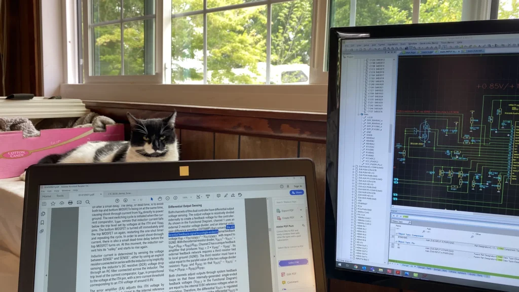 Black-and-white cat lounging on pink bed beside laptop and monitor displaying PCB schematics in sunny room.