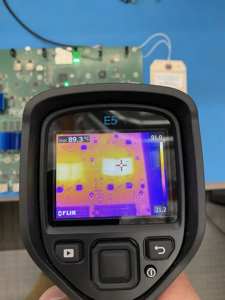 Handheld FLIR thermal camera displaying heat map of PCB with max 89.3°C hotspot.