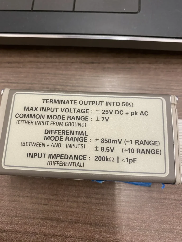 Gray BNC differential amplifier module with electrical specs label on wooden desk.