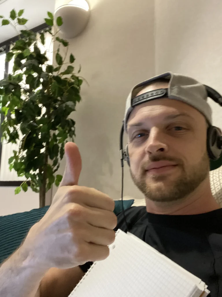 Man in gray cap and headphones giving thumbs up beside potted plant and notebook.