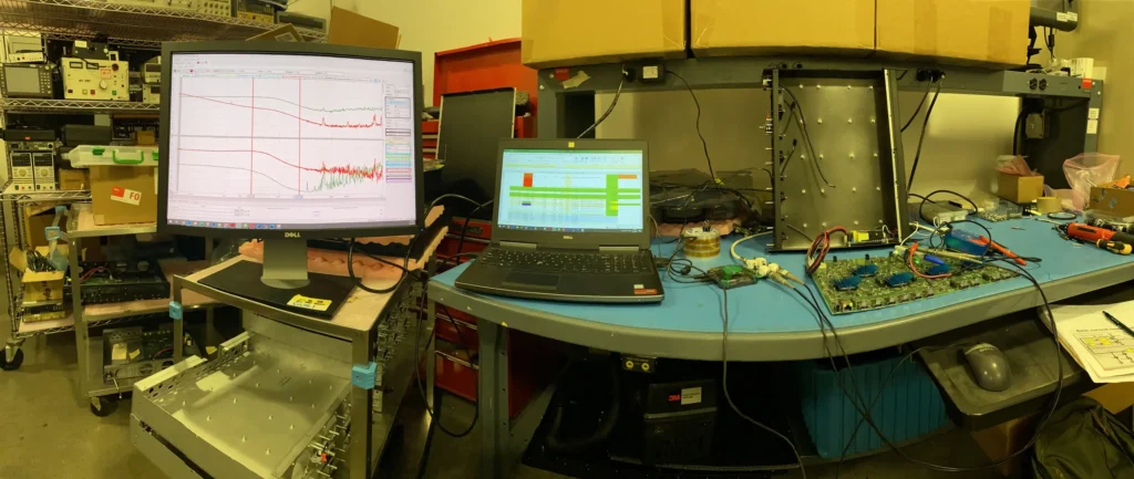 Electronics workbench with oscilloscope displaying waveforms, laptops, circuit boards, tools, and lab equipment.