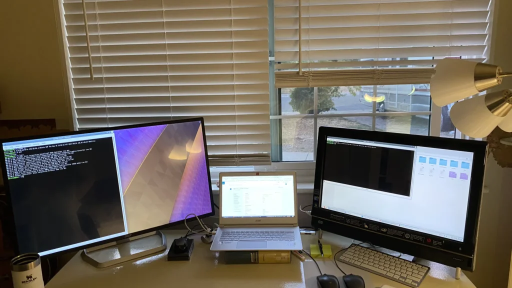 Dual monitors displaying terminal and software interfaces flanking a laptop on a desk by a window with blinds.