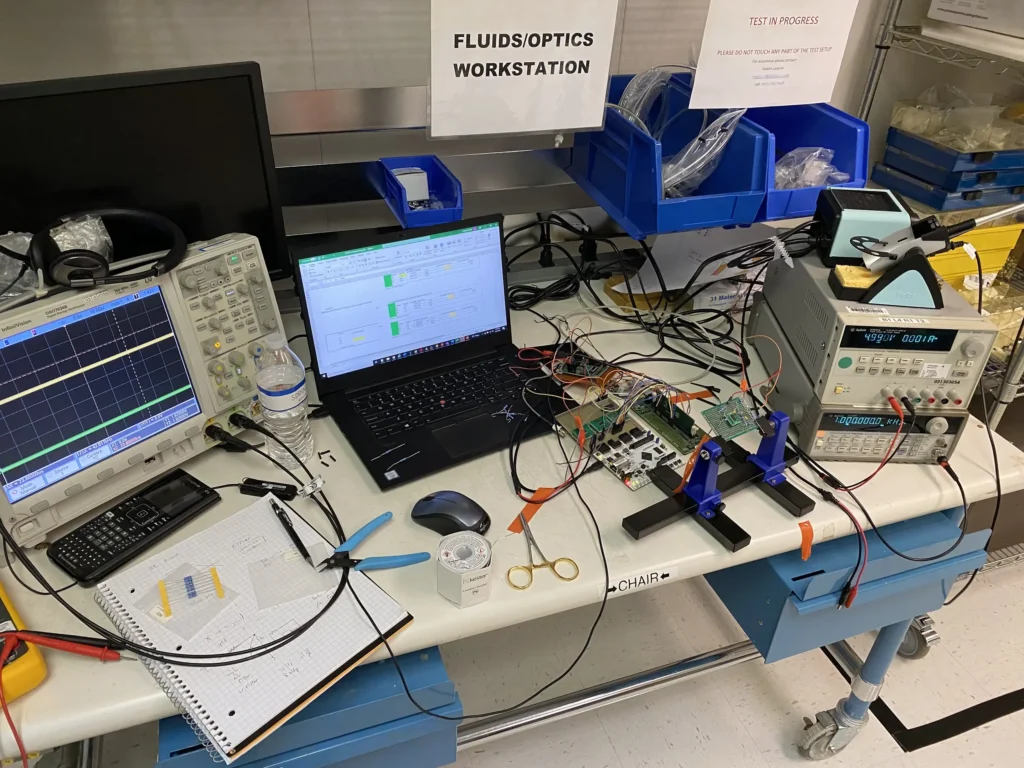 Cluttered fluids/optics workstation with oscilloscopes, multimeters, laptops, circuit boards, and test equipment on a lab bench.
