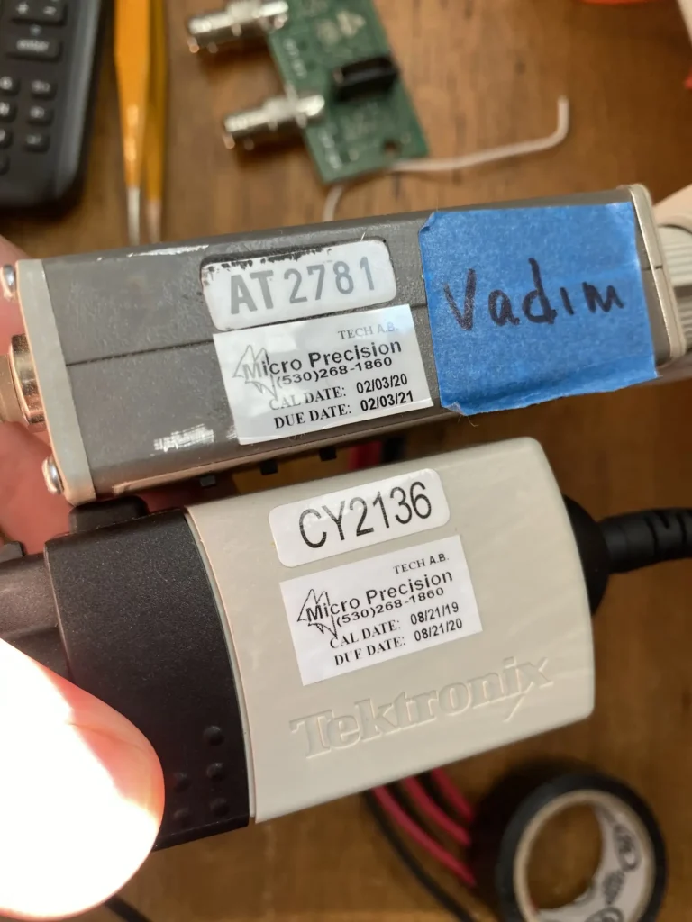 Two labeled Micro Precision calibration adapters (AT2781 and CY2136) held in hands next to Tektronix probe on tech lab bench.