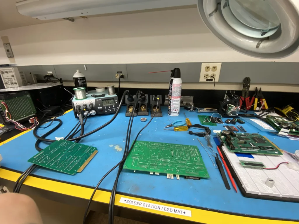 Electronics workbench with soldering station, tools, green circuit boards, and ESD mat.