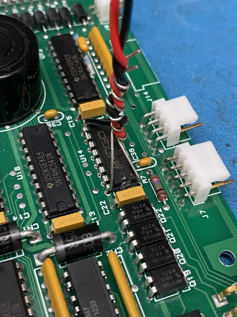 Close-up of a green PCB with black IC chips, yellow capacitors, red and yellow wires, and white connectors on a blue surface.
