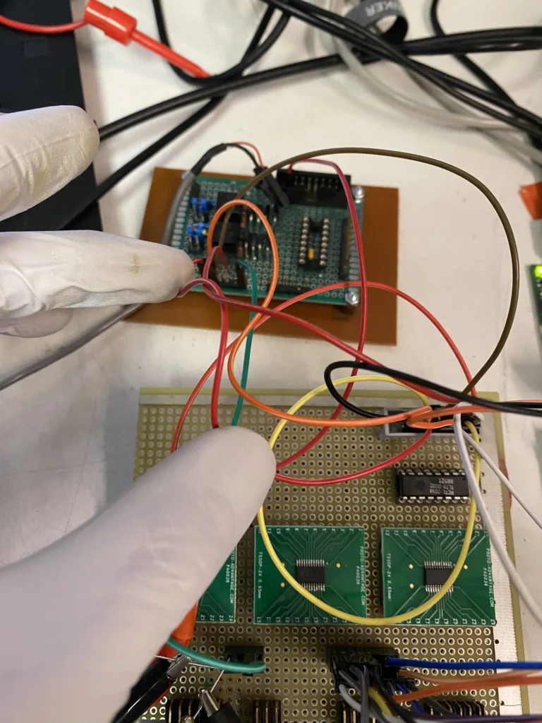 Gloved hands wiring colorful components and ICs on a green protoboard amid tangled cables.