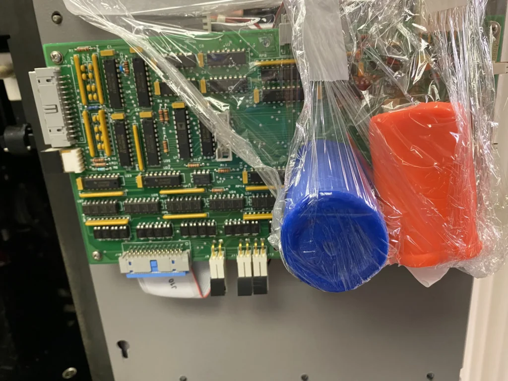 Green circuit board with IC chips and connectors wrapped in plastic alongside blue and orange playdough.
