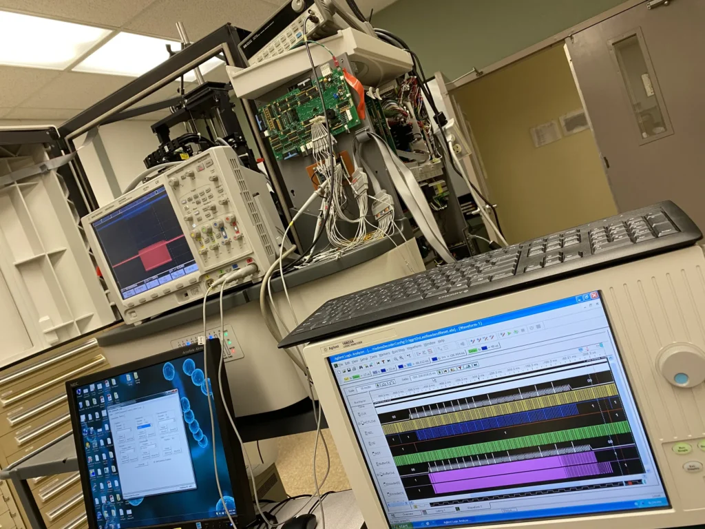 Electronics lab bench with oscilloscope displaying waveform, logic analyzer screen with multi-channel traces, and interconnected circuit boards.