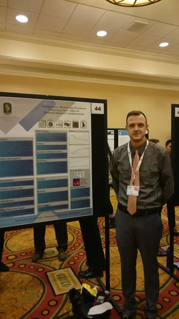 Man in gray shirt and lanyard standing beside poster on "Temperature Monitoring Footwear for Early Detection of Diabetes-Related Complications" at conference.