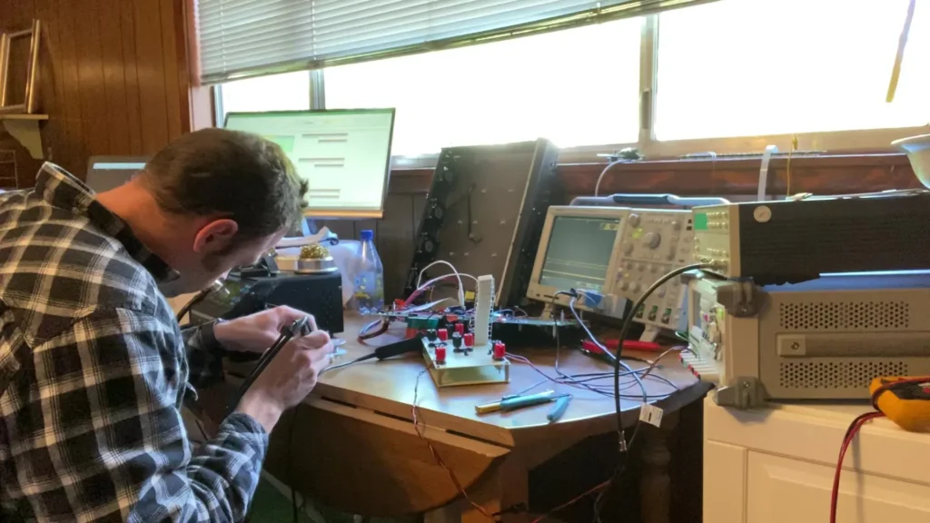 Man in plaid shirt soldering electronics on workbench with oscilloscopes, multimeter, and computer monitors.