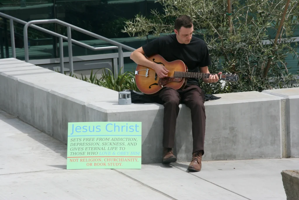 Man in black shirt playing archtop guitar on concrete ledge beside sign reading "Jesus Christ sets free from addiction and gives eternal life."