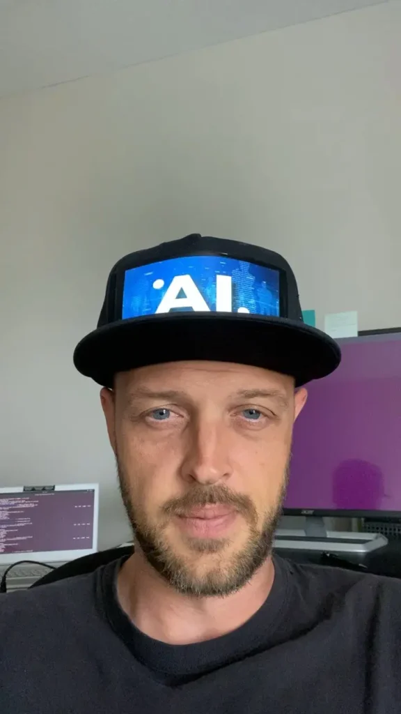 Man with goatee wearing black cap with glowing blue "AI" display, sitting before computer screens.